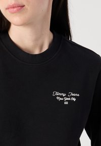 Et nært bilde av en persons nakke og hake, iført en svart hettegenser med teksten "Tommy Jeans New York City" i hvit brodering.