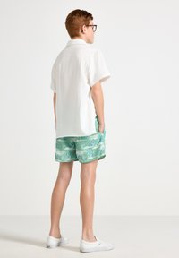Camicetta bianca a maniche corte con texture, pantaloni corti verdi con fantasia di palme e scarpe bianche slip-on. Bambino visto da dietro.