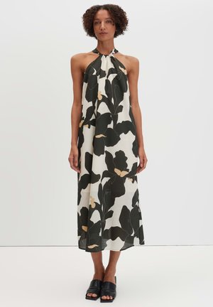 Femme portant une robe midi sans manches col halter avec un grand imprimé floral noir et beige, associée à des chaussures noires ouvertes à l'avant et sans lacets.