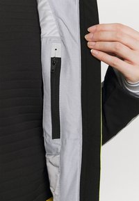 Main tenant une veste noire ouverte montrant la doublure intérieure grise avec une poche zippée verticale et un détail en tissu noir.