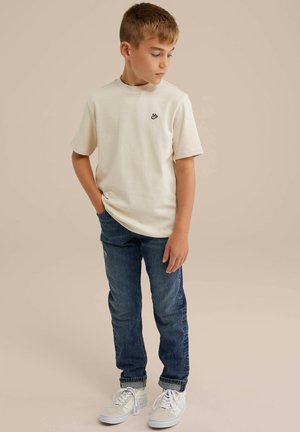 Beige Kurzarm-T-Shirt mit Rundhalsausschnitt und schwarzem Logo auf der Brust, kombiniert mit blauen Jeans und weißen Sneakern.