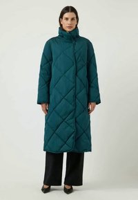 Teal quilted long coat with diamonds pattern, high collar, full-length sleeves, and a straight silhouette. Worn over black pants and shoes.