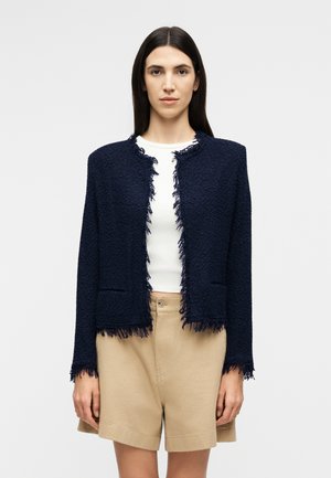 Γυναίκα με μακριά σκουρόχρωμα μαλλιά, ντυμένη με μπλε υφασμάτινο ανοιχτό σακάκι με κρόσσια πάνω από λευκό τοπ και μπεζ σορτς.