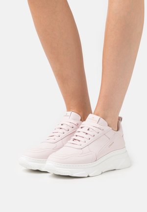 Baskets légères roses pâles à lacets avec semelles blanches épaisses portées pieds nus sur un fond blanc uni.