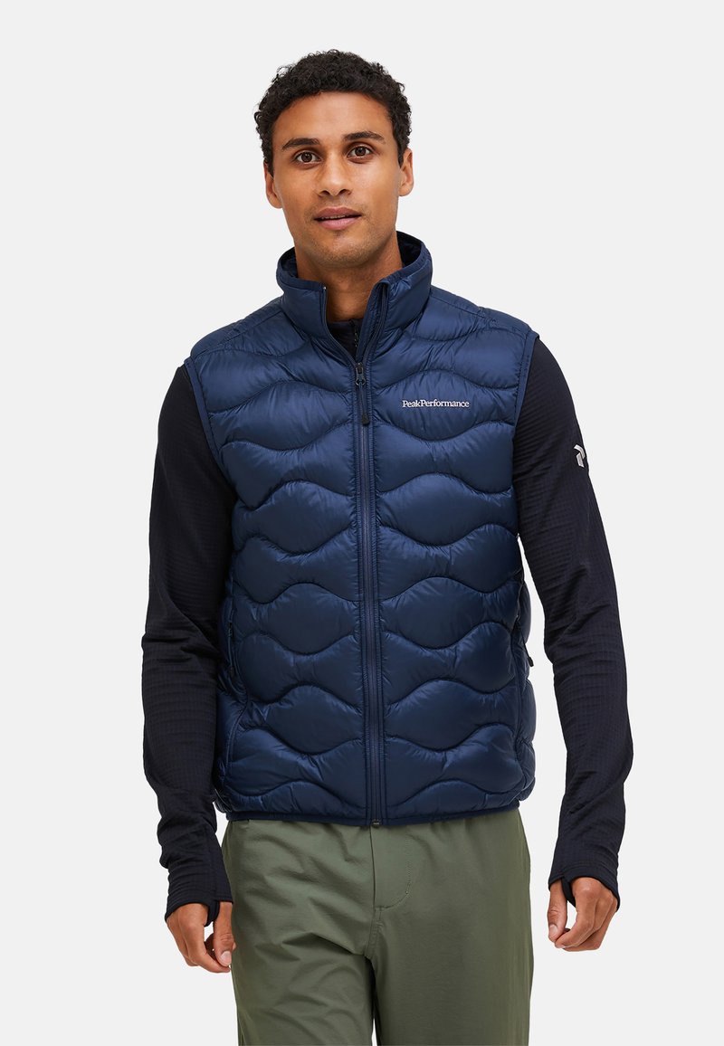 Navy quilted puffer vest with high collar, zip front, and logo. Worn over a long-sleeve shirt, paired with olive green pants. Lightweight material.