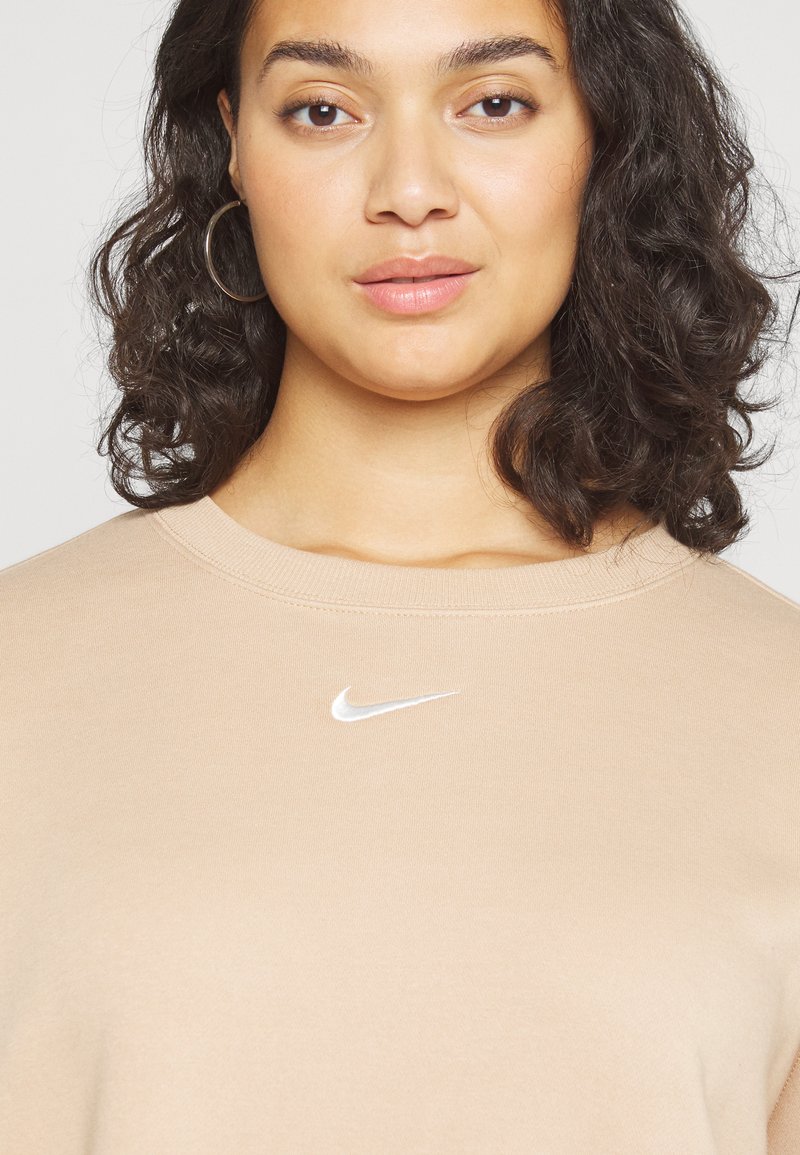 Sweatshirt beige clair avec un col rond, arborant un logo Nike blanc sur la poitrine. Le tissu semble doux et lisse. Cheveux bouclés visibles.