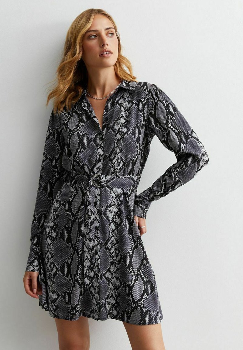 Frau mit blonden Haaren, die ein langärmeliges, mit Gürtel versehenes, schwarz-grau gemustertes Kleid im Schlangenmuster trägt, steht vor einer schlichten hellen Wand.