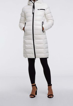 Femme porte un manteau matelassé blanc long avec une fermeture éclair noire, des leggings noirs et des sandales noires à talons hauts, debout les mains dans les poches.