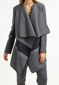 Mujer vestida con un abrigo gris de frente abierto y amplias solapas sobre una blusa estampada y pantalones gris a juego, sobre un fondo blanco.