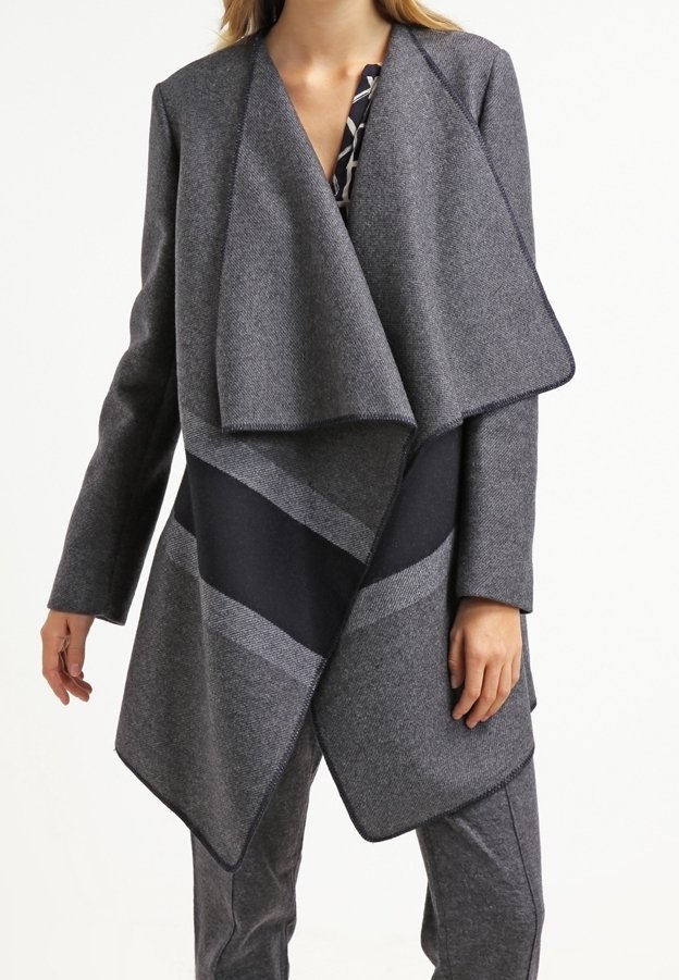 Mujer vestida con un abrigo gris de frente abierto y amplias solapas sobre una blusa estampada y pantalones gris a juego, sobre un fondo blanco.