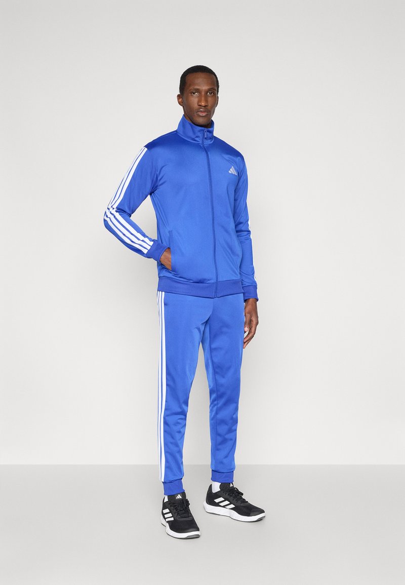 Blauer Trainingsanzug mit einer zip-up Jacke und schmalen Hosen, verziert mit weißen Streifen entlang der Ärmel und Beine, kombiniert mit schwarzen Turnschuhen.