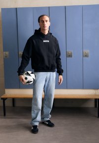 Čierna mikina s nápisom "JUVENTUS", svetlomodré džínsy, čierne tenisky, drží čierno-biely vzorovaný futbalový loptu, stojí pred skriňami.