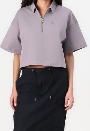 Vrouw draagt een lavendelkleurig cropped poloshirt met rits en een zwarte hoge taille rok met trekkoorden en zakken.