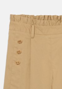 Pantalons beiges avec une taille froncée, ornés de quatre boutons décoratifs en or sur le côté et d'un tissu lisse et texturé.