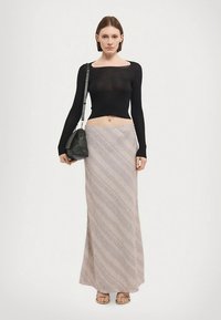 Nő áll, fekete, hosszú ujjú, rövidített felsőt, bézs mintás maxi szoknyát, arany szandált visel, és egy sötétzöld válltáskát tart.