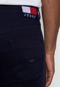 Jeans de mezclilla azul marino con un bolsillo trasero, que cuentan con una pequeña etiqueta de marca con acentos en rojo, blanco y azul y un parche textil en la cinturilla.