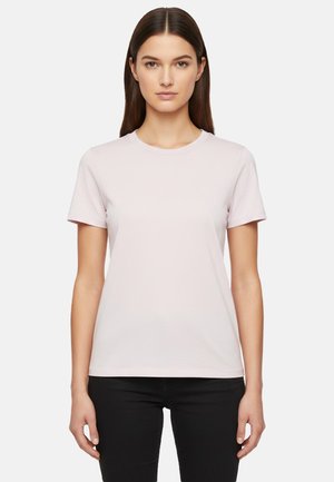 Giovane donna con capelli scuri lisci che indossa una maglietta rosa chiaro a maniche corte semplice e pantaloni neri, in piedi davanti a uno sfondo bianco.