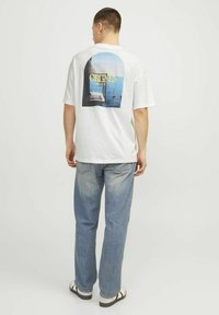 Hvid bomulds t-shirt med et grafisk tryk på ryggen, der viser blåt hav og sejlbåde, parret med lyseblå denim jeans og sandaler.