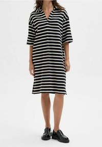 Femme portant une robe noire et blanche à rayures horizontales, longueur genou, à manches courtes et des mocassins noirs brillants, se tenant sur un fond blanc.