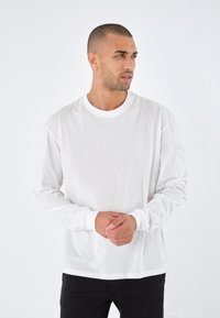 Witte katoenen blouse met lange mouwen, een ronde halslijn, een ontspannen pasvorm en geribbelde manchetten, gedragen met zwarte broek voor een effen achtergrond.