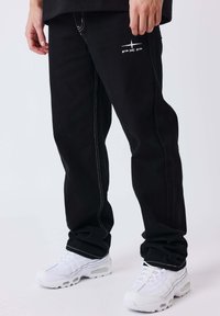 Persoon draagt zwarte broek met wit logo op dij en witte sportschoenen, staand tegen een effen achtergrond.