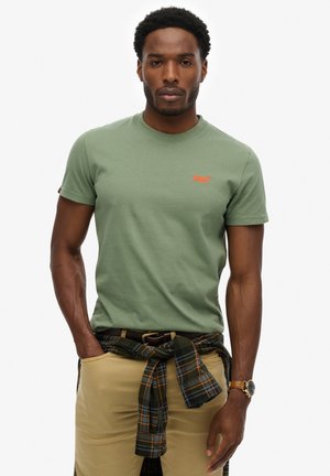 Man die een groen T-shirt met korte mouwen en een klein oranje logo draagt, beige broek, geruite blouse om de taille gestrikt en een polshorloge, staand tegen een witte achtergrond.