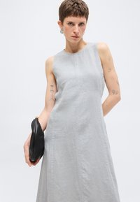 Γυναίκα με κοντά μαλλιά που φοράει αμάνικο γκρι φόρεμα, κρατάει μαύρη τσάντα φάκελο και δείχνει διάφορα τατουάζ στα μπράτσα της.