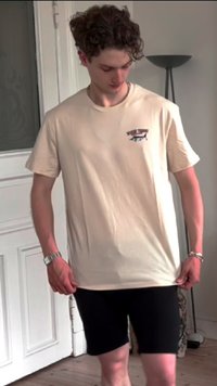 Beige bomulds t-shirt med korte ærmer, der har en lille grafisk detalje på venstre bryst. Sat sammen med sorte shorts, står indendørs.