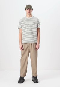 Cămașă cu mânecă scurtă, în dungi, de culoare crem și verde închis, asortată cu pantaloni elastici bej și adidași negri. Modelul poartă o șapcă verde închis.