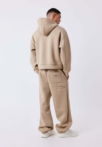 Man in beige oversized hoodie en bijpassende joggingbroek met witte sneakers, met handen in de zakken, de andere kant op kijkend.