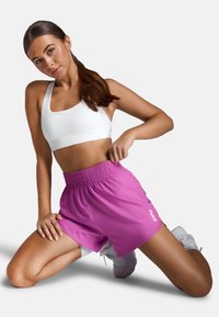 Lyserøde idrætsshorts med elastisk talje og hvidt logo; parret med en hvid sports-bh. Model er på knæ.