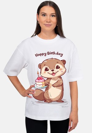 Junge Frau trägt ein weißes T-Shirt mit dem Schriftzug "Happy Birthday" und einem cartoonartigen Otter, der einen Kuchen mit brennender Kerze hält.