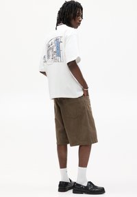 Witte t-shirt met korte mouwen en een graphic op de achterkant, gecombineerd met bruine corduroy shorts. Zwarte schoenen en witte crew sokken maken de outfit compleet.