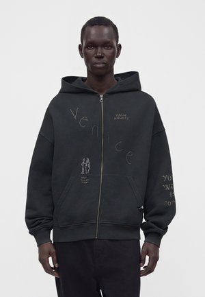 Mand iført en sort lynlåshættetrøje med teksten "Venice", små hvide tegninger og "Palm Angels" logo på brystet mod en ensfarvet lys baggrund.