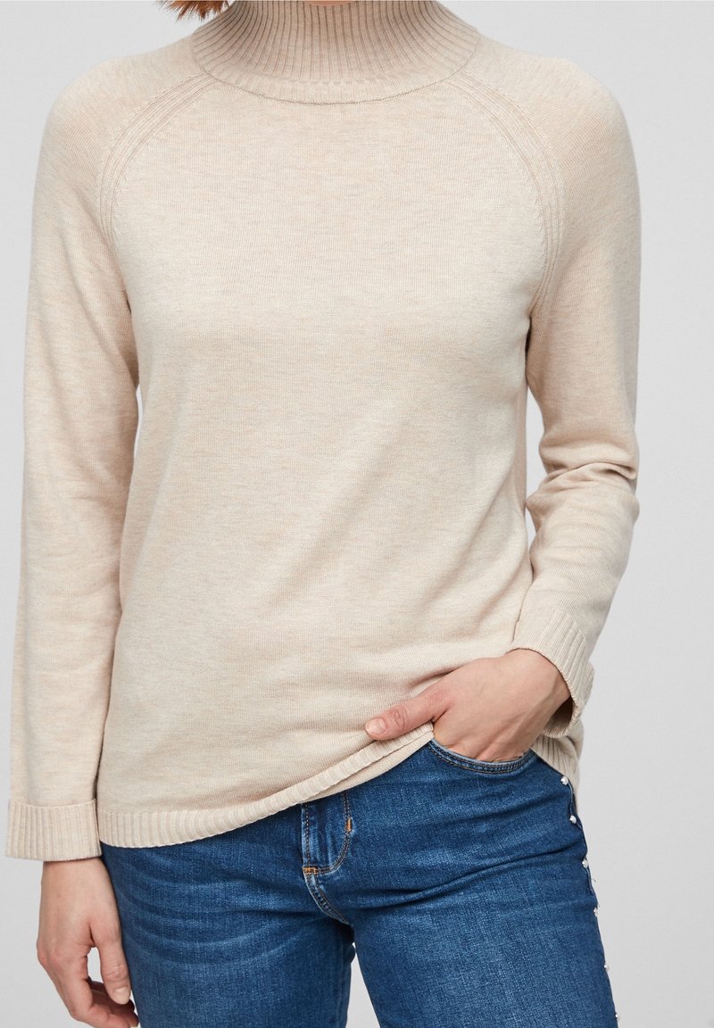 Personne portant un pull à col roulé beige et un jean bleu, main dans la poche, sur fond uni.