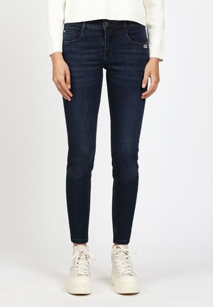 Person wearing dark blue skinny jeans and white high-top sneakers, standing against a plain light background.