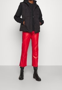 Veste matelassée noire avec capuche portée sur une chemise noire, associée à un pantalon en cuir rouge et des bottines noires. Textures lisses et coupe ajustée.