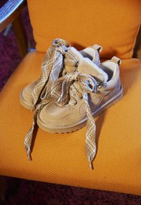 Light beige sneakers feature a textured suede upper, distinctive patterned laces, and a cushioned interior. Placed on an orange chair.
