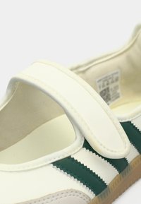 Άσπρο δερμάτινο παπούτσι με πράσινες λωρίδες και ιμάντα Velcro, με στρογγυλή μύτη και υφασμένη ελαστική σόλα. Το εσωτερικό επένδυμα είναι απαλό μπεζ.