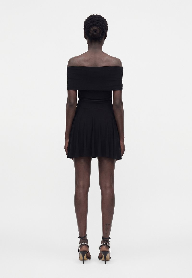 Femme à la peau foncée et aux cheveux attachés, portant une robe noire courte, épaules dénudées, et des sandales à talons hauts, debout de dos, tournée vers l'avant.