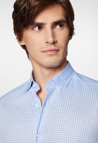 Jasnoniebieska koszula z guzikami, z małym geometrycznym wzorem, standardowym kołnierzykiem i gładką fakturą materiału, podkreślająca subtelne detale wzornicze.