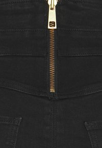 Musta farkkukangas, jossa edessä kultainen vetoketjun vetolaite. Huomattava ompelu, kaksi etutaskua ja sileä pinta.