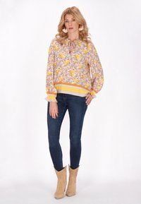 Blusa floreale con motivi gialli e rosa, maniche lunghe, colletto con laccetto e polsini volant; abbinata a jeans skinny scuri e stivali da cowboy color sabbia.