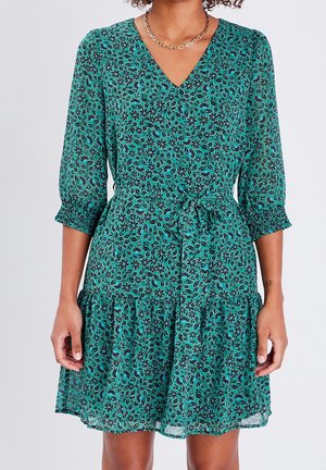 Femme portant une robe fleurie bleu sarcelle à manches trois-quarts, col en V et ceinture nouée à la taille, debout devant un fond clair uni.
