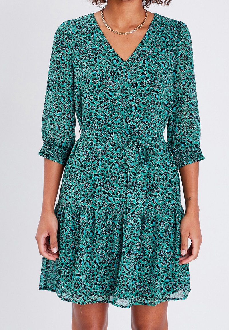Femme portant une robe fleurie bleu sarcelle à manches trois-quarts, col en V et ceinture nouée à la taille, debout devant un fond clair uni.