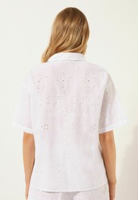 Camicetta bianca a maniche corte con ricami floreali e dettagli ad occhio di pernice. Design con colletto e una leggerissima trasparenza.