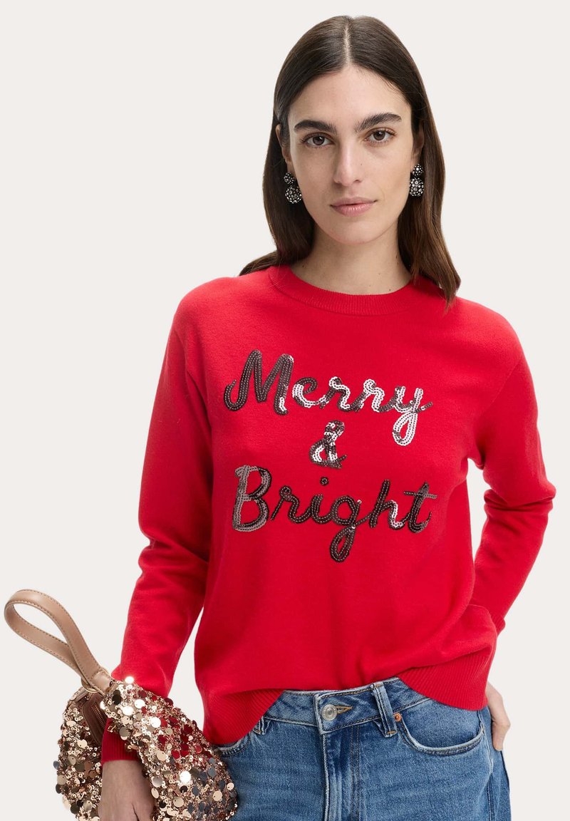 Maglione rosso con "Merry & Bright" in paillettes argento. Collo rotondo, maniche lunghe, abbinato a una borsa in paillettes. Jeans in denim sotto.