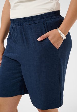 Personne portant un short bleu marine texturé avec une taille élastique et la main dans la poche droite, bracelet argenté visible au poignet.