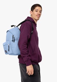 Modrá batoh s několika zipy a kapsami; nošený s fialovou mikinou a černými kalhotami. Má nastavitelné popruhy a strukturovaný tvar.