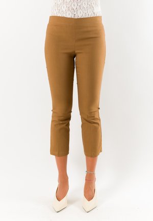 Donna che indossa pantaloni marroni corti, scarpe bianche con tacchi alti e una maglia bianca di pizzo, in piedi davanti a uno sfondo chiaro e uniforme.